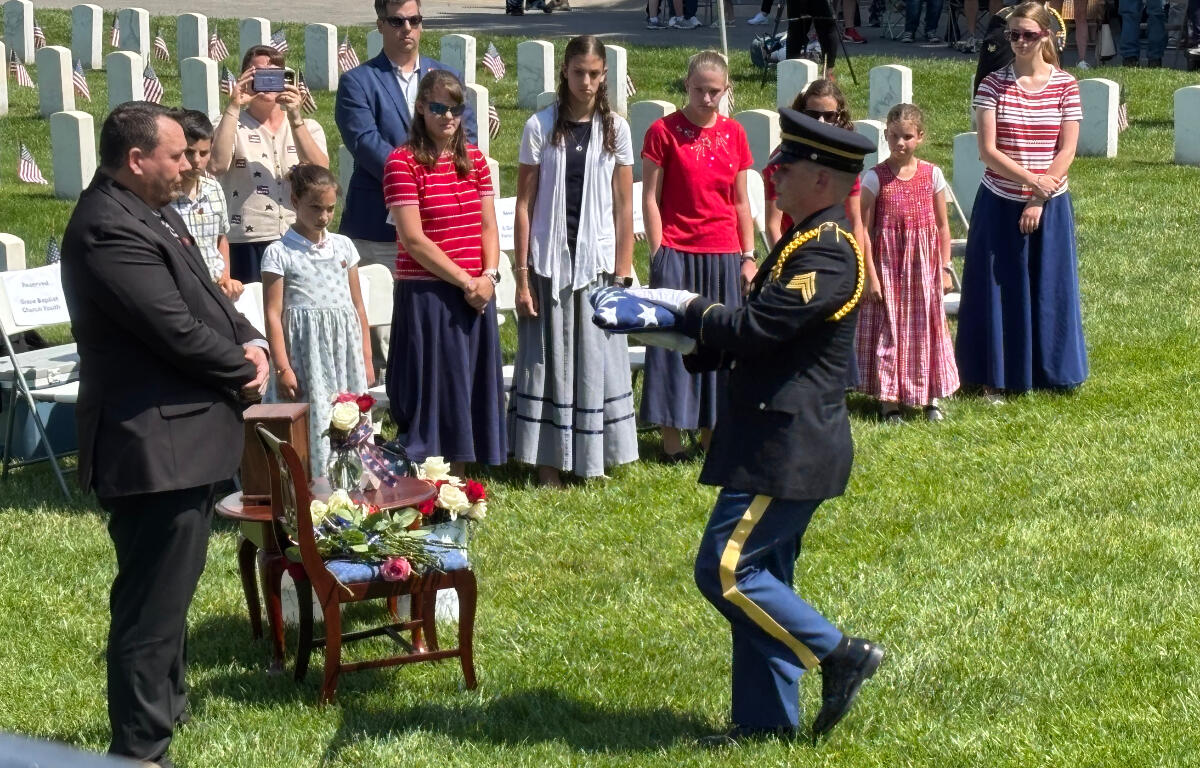 Military honor guard presenting folded American flag at gravesite as attendees watch during a memorial ceremony on a sunny day.