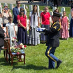 Military honor guard presenting folded American flag at gravesite as attendees watch during a memorial ceremony on a sunny day.