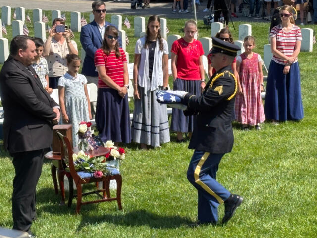 Military honor guard presenting folded American flag at gravesite as attendees watch during a memorial ceremony on a sunny day.