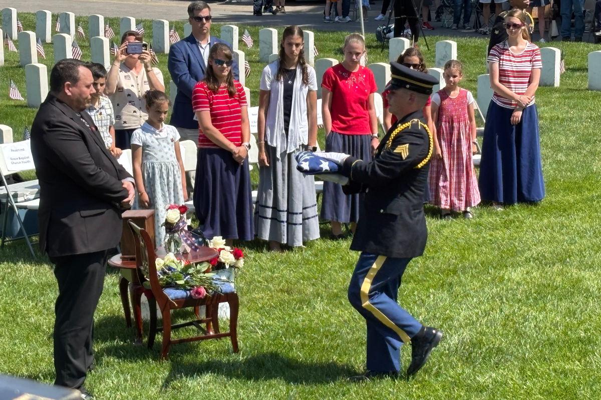 Military honor guard presenting folded American flag at gravesite as attendees watch during a memorial ceremony on a sunny day.