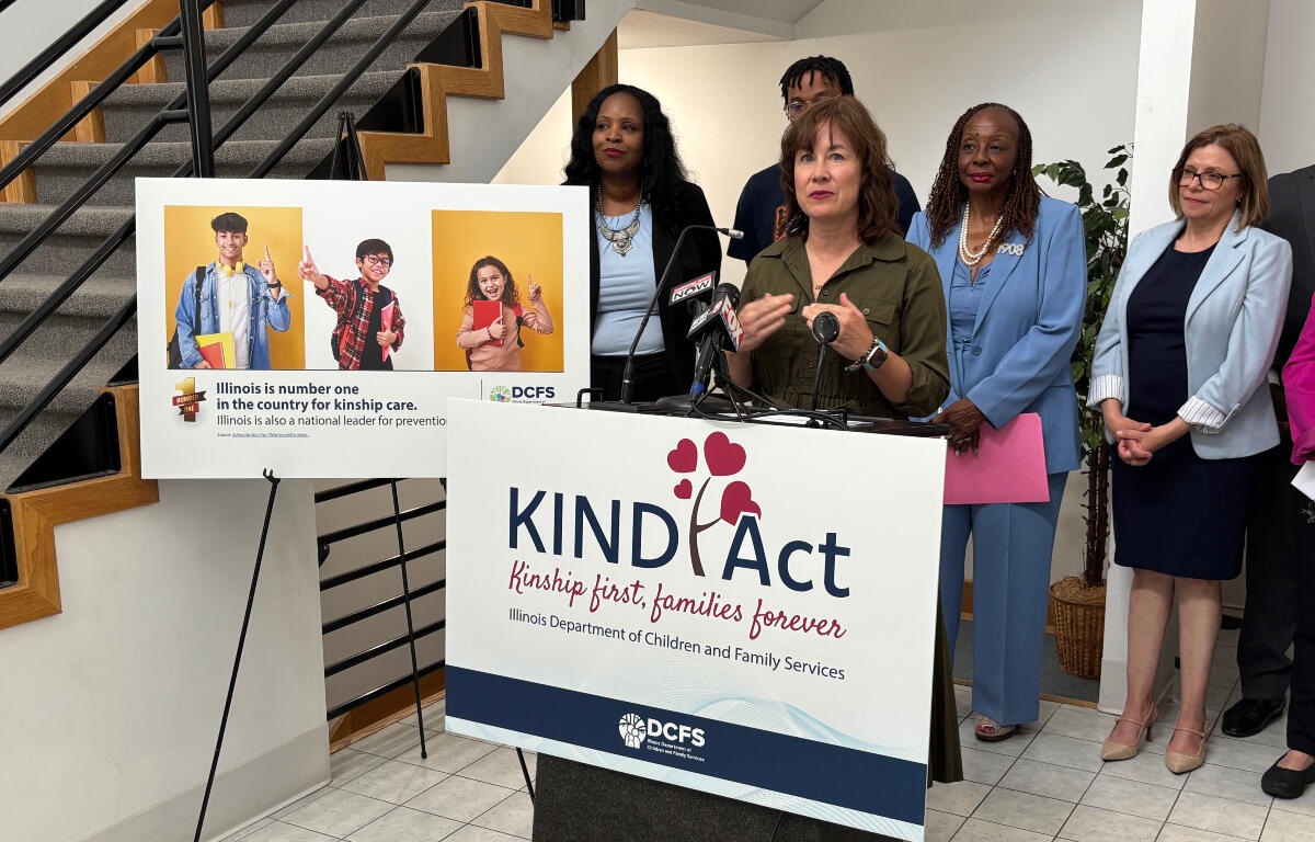 Speaker at a KIND Act press conference, with a podium and a banner that reads KIND Act: Kinship first, families forever, while diverse officials stand nearby and a poster with three kids is displayed to the left.