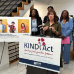 Speaker at a KIND Act press conference, with a podium and a banner that reads KIND Act: Kinship first, families forever, while diverse officials stand nearby and a poster with three kids is displayed to the left.
