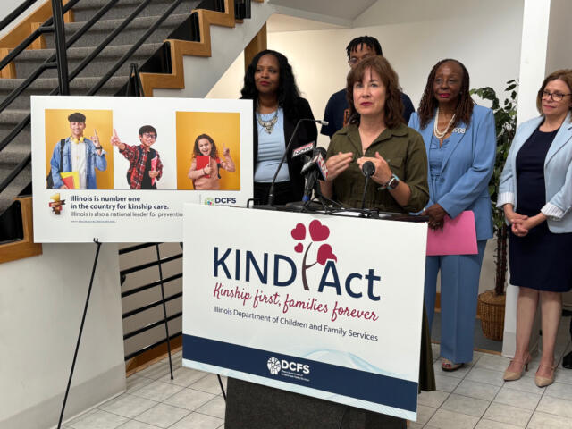 Speaker at a KIND Act press conference, with a podium and a banner that reads KIND Act: Kinship first, families forever, while diverse officials stand nearby and a poster with three kids is displayed to the left.