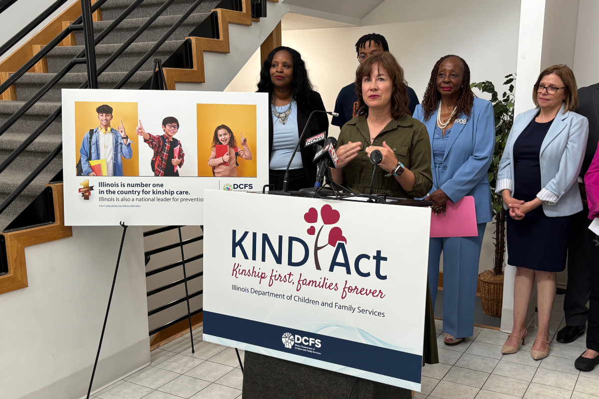 Speaker at a KIND Act press conference, with a podium and a banner that reads KIND Act: Kinship first, families forever, while diverse officials stand nearby and a poster with three kids is displayed to the left.