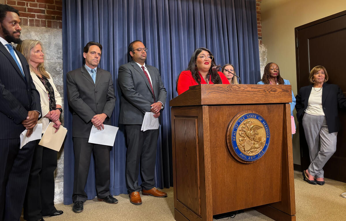 Woman in red at a lectern delivering remarks; diverse officials stand behind her at a press conference with a blue curtain backdrop.