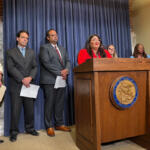 Woman in red at a lectern delivering remarks; diverse officials stand behind her at a press conference with a blue curtain backdrop.