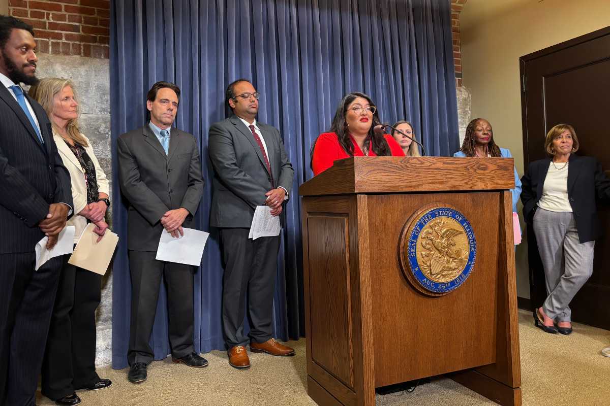 Woman in red at a lectern delivering remarks; diverse officials stand behind her at a press conference with a blue curtain backdrop.