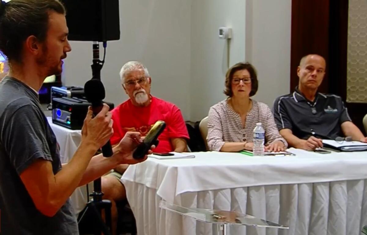 A man with a microphone speaks to a panel of three seated at a long table during a meeting or presentation.