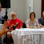 A man with a microphone speaks to a panel of three seated at a long table during a meeting or presentation.