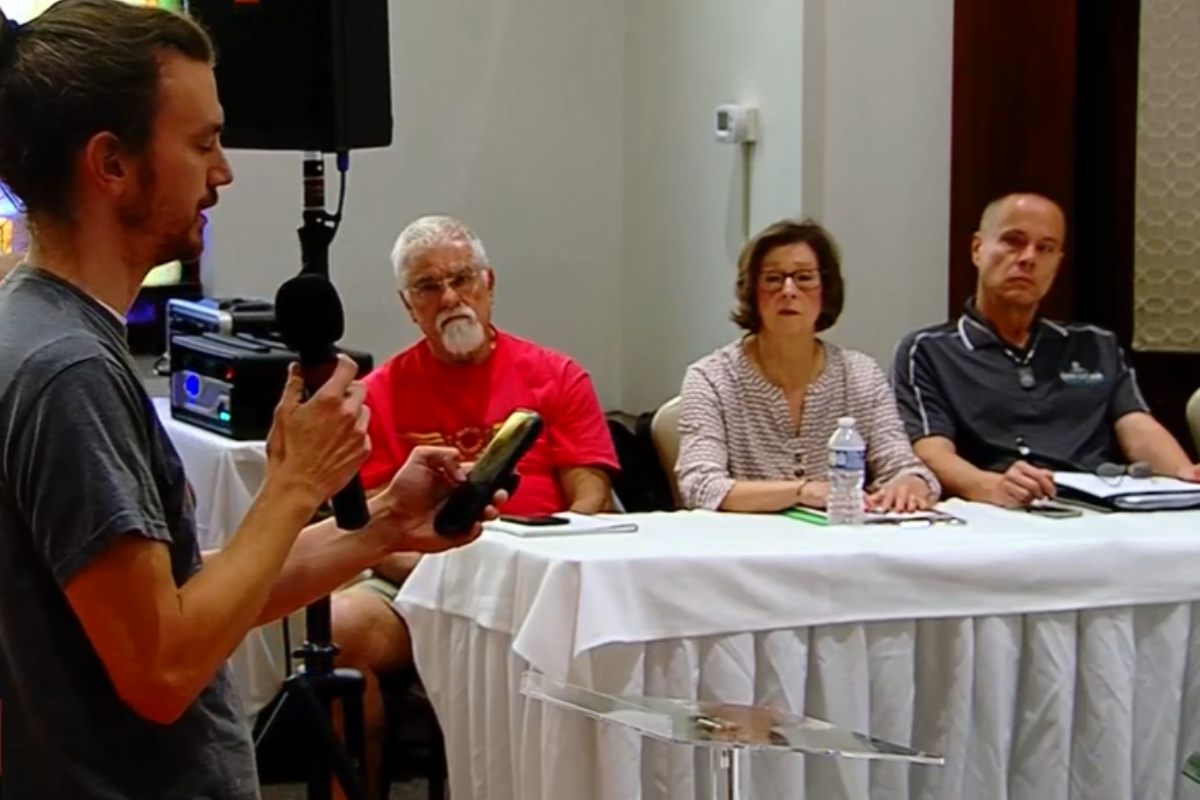 A man with a microphone speaks to a panel of three seated at a long table during a meeting or presentation.