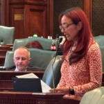 Woman with long red hair and glasses speaks at a microphone in a wood-paneled legislative chamber close to a desk and papers.