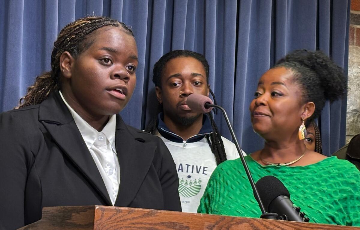 Young woman in a black suit speaks at a wooden podium, with a man behind and a smiling woman in a green top nearby in front of blue curtains.