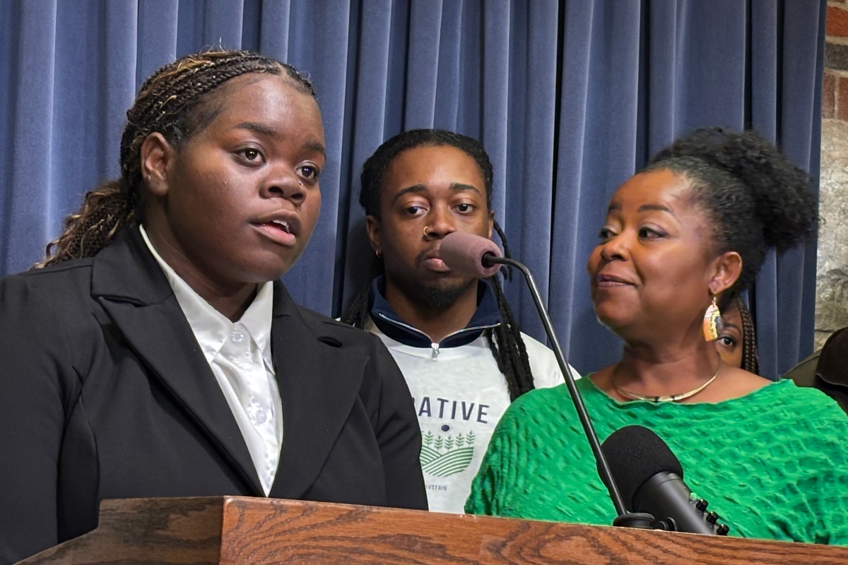 Young woman in a black suit speaks at a wooden podium, with a man behind and a smiling woman in a green top nearby in front of blue curtains.