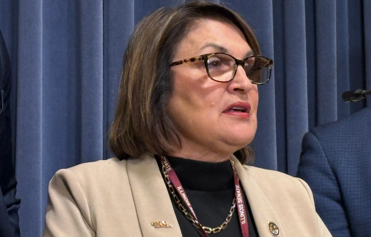 Woman with glasses and a beige blazer speaks at a podium, Illinois Senate lanyard visible in front of blue curtains.