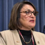 Woman with glasses and a beige blazer speaks at a podium, Illinois Senate lanyard visible in front of blue curtains.