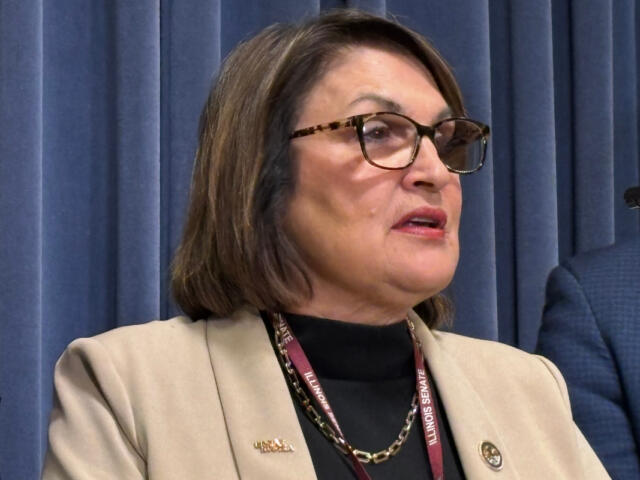 Woman with glasses and a beige blazer speaks at a podium, Illinois Senate lanyard visible in front of blue curtains.