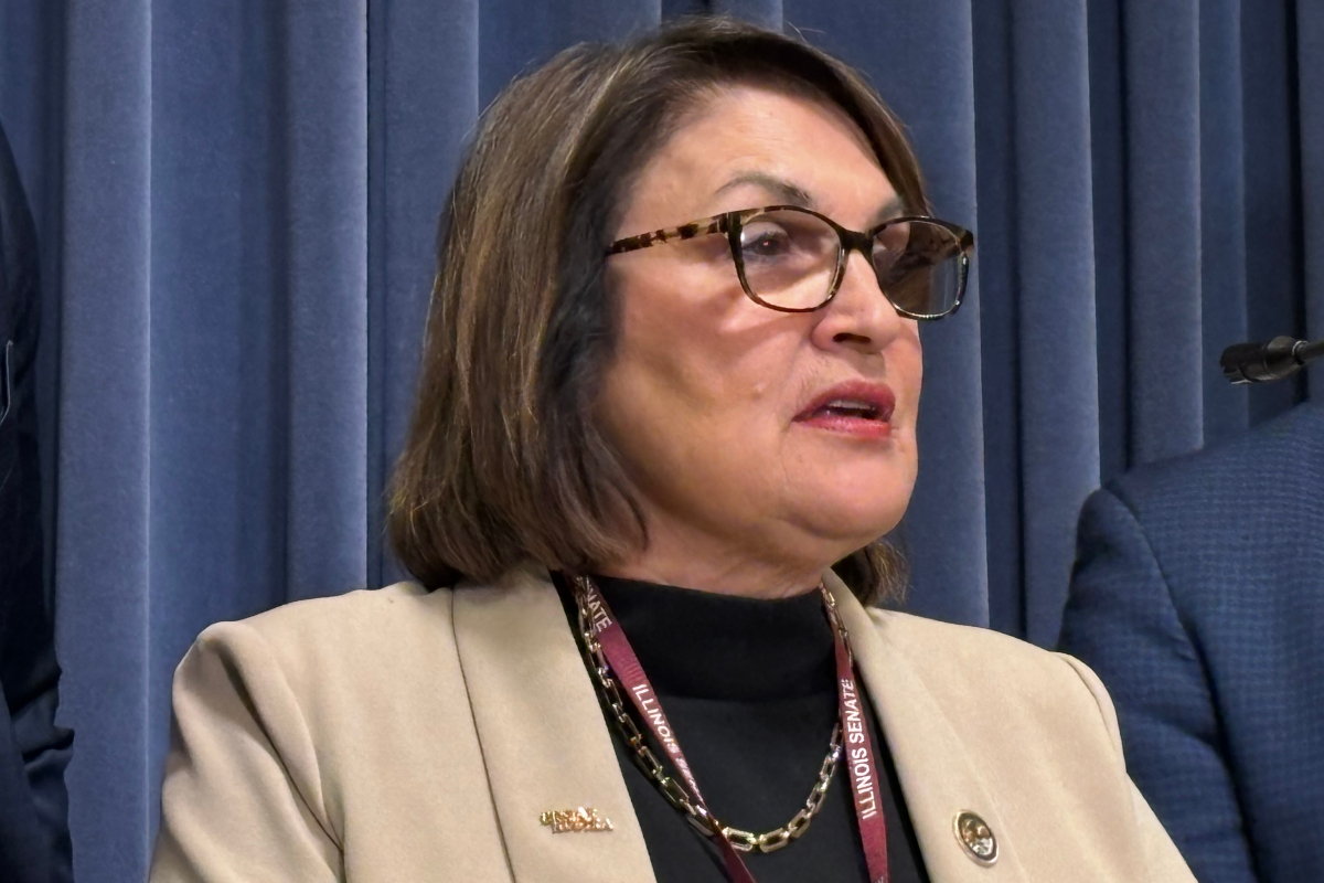 Woman with glasses and a beige blazer speaks at a podium, Illinois Senate lanyard visible in front of blue curtains.