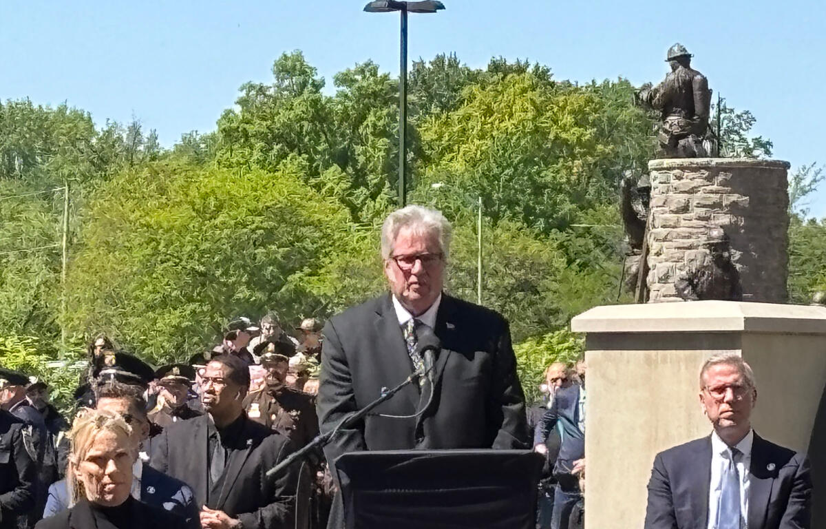 Man in a dark suit speaks at a podium with microphones during an outdoor ceremony, a crowd and statue in the background, sunny day.