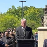 Man in a dark suit speaks at a podium with microphones during an outdoor ceremony, a crowd and statue in the background, sunny day.