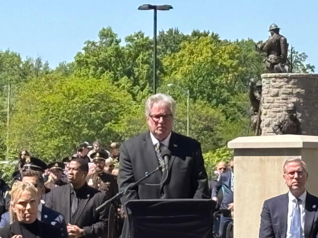 Man in a dark suit speaks at a podium with microphones during an outdoor ceremony, a crowd and statue in the background, sunny day.