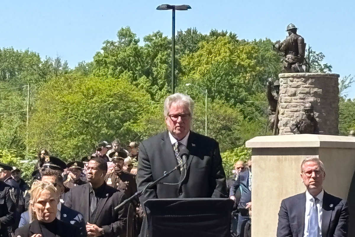 Man in a dark suit speaks at a podium with microphones during an outdoor ceremony, a crowd and statue in the background, sunny day.