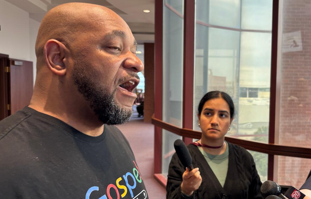 Man with a shaved head and beard speaks to reporters in a hallway while a young female reporter holds a handheld microphone nearby.