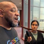 Man with a shaved head and beard speaks to reporters in a hallway while a young female reporter holds a handheld microphone nearby.