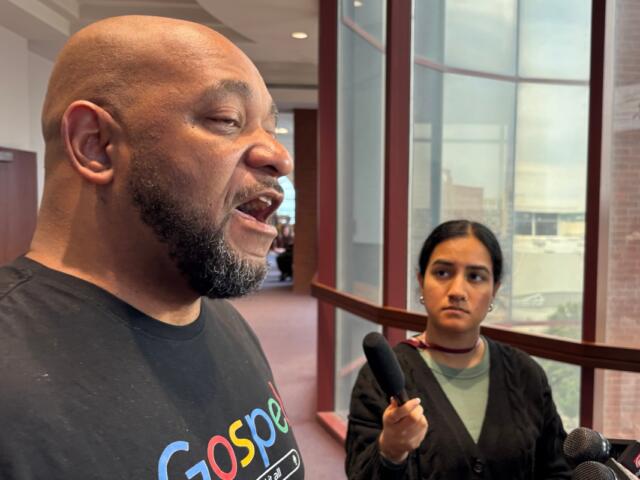 Man with a shaved head and beard speaks to reporters in a hallway while a young female reporter holds a handheld microphone nearby.