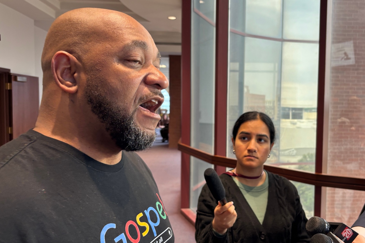 Man with a shaved head and beard speaks to reporters in a hallway while a young female reporter holds a handheld microphone nearby.