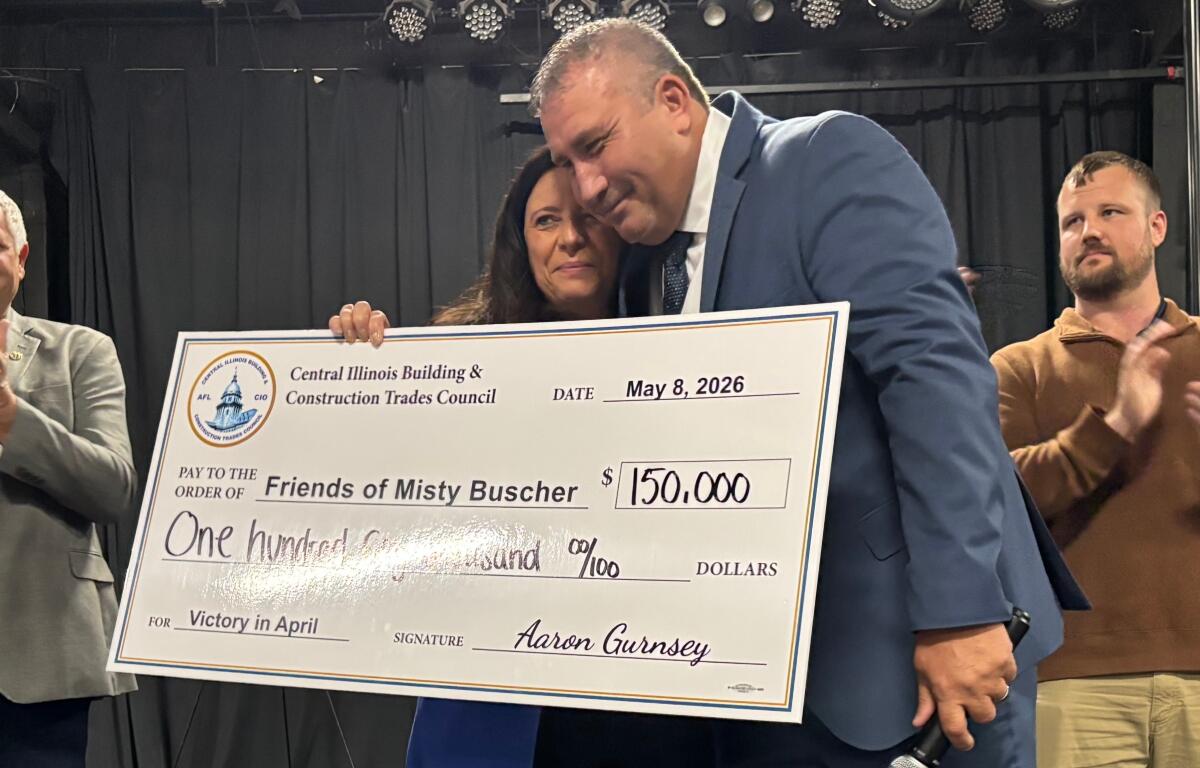 Two people in formal attire hold a large ceremonial check from the Central Illinois Building & Construction Trades Council, dated May 8, 2026, for $150,000 to 'Friends of Misty Busher' with signatures visible, as another man applauds in the background.