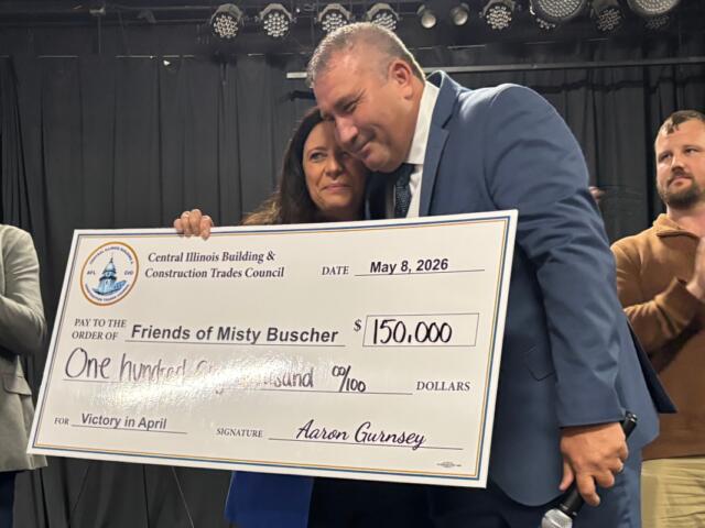 Two people in formal attire hold a large ceremonial check from the Central Illinois Building & Construction Trades Council, dated May 8, 2026, for $150,000 to 'Friends of Misty Busher' with signatures visible, as another man applauds in the background.