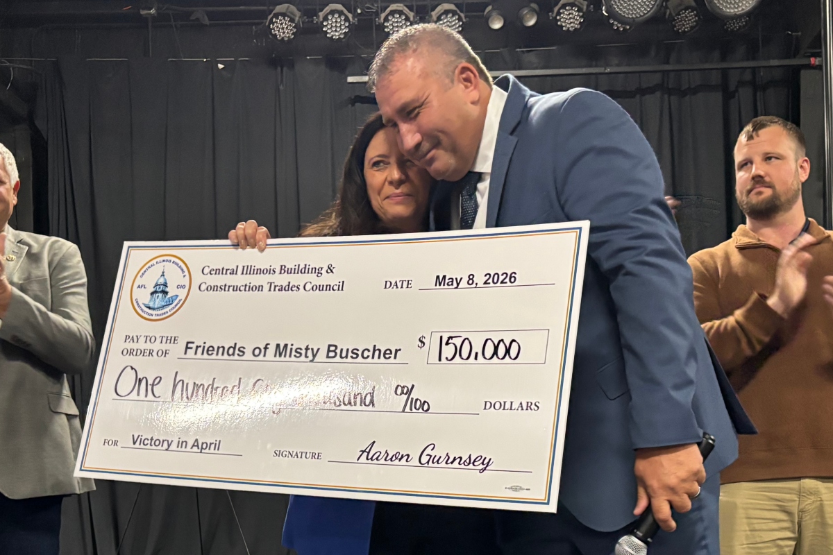 Two people in formal attire hold a large ceremonial check from the Central Illinois Building & Construction Trades Council, dated May 8, 2026, for $150,000 to 'Friends of Misty Busher' with signatures visible, as another man applauds in the background.