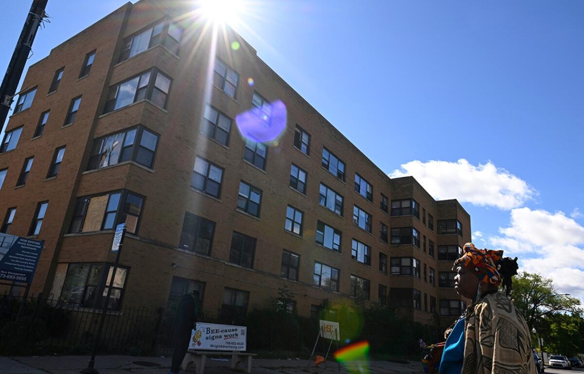 Brick apartment building angled under bright sun with lens flare; a person with a colorful headwrap stands on the sidewalk to the right.