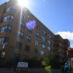 Brick apartment building angled under bright sun with lens flare; a person with a colorful headwrap stands on the sidewalk to the right.