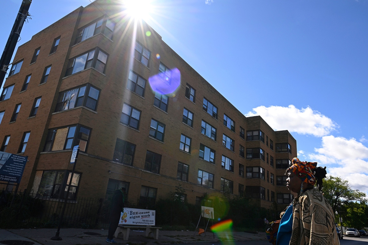 Brick apartment building angled under bright sun with lens flare; a person with a colorful headwrap stands on the sidewalk to the right.