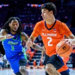 Two college basketball players sprint toward the hoop during a fast break; Illinois in orange and FGCU in blue facing off on a crowded court.