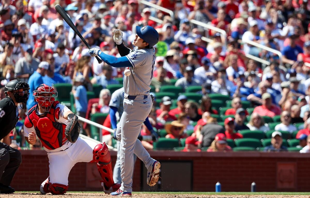 Baseball batter in gray swings at a pitch as a red‑gear catcher and umpire look on with a crowded stadium in the background, sunny day.