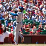 Baseball batter in gray swings at a pitch as a red‑gear catcher and umpire look on with a crowded stadium in the background, sunny day.