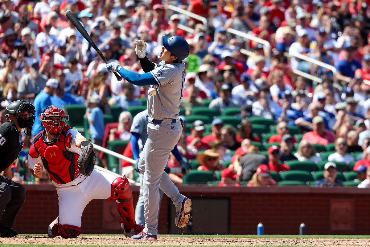 Baseball batter in gray swings at a pitch as a red‑gear catcher and umpire look on with a crowded stadium in the background, sunny day.