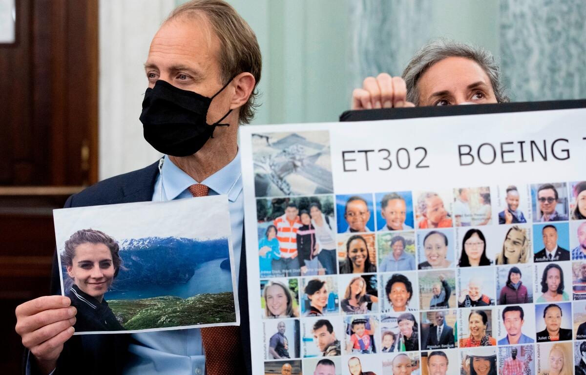 Man in a suit and black mask holds a landscape photo next to a large poster labeled ET302 Boeing with many portrait thumbnails.