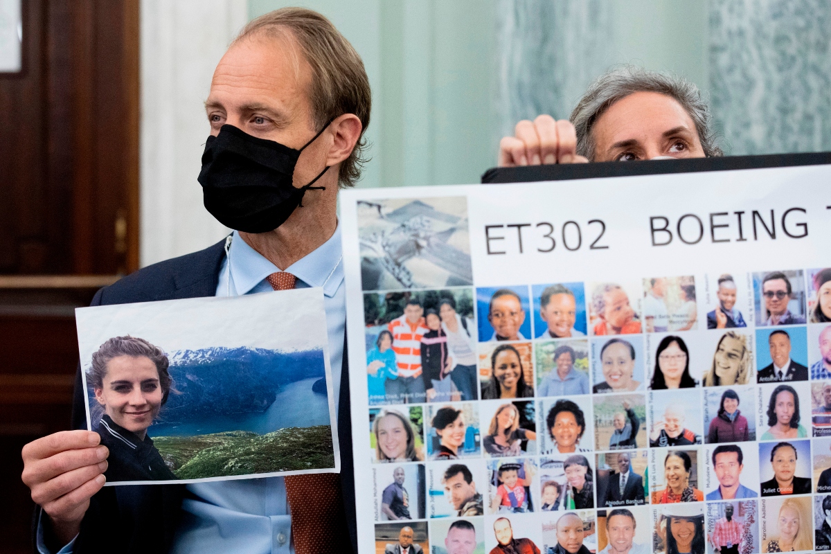 Man in a suit and black mask holds a landscape photo next to a large poster labeled ET302 Boeing with many portrait thumbnails.
