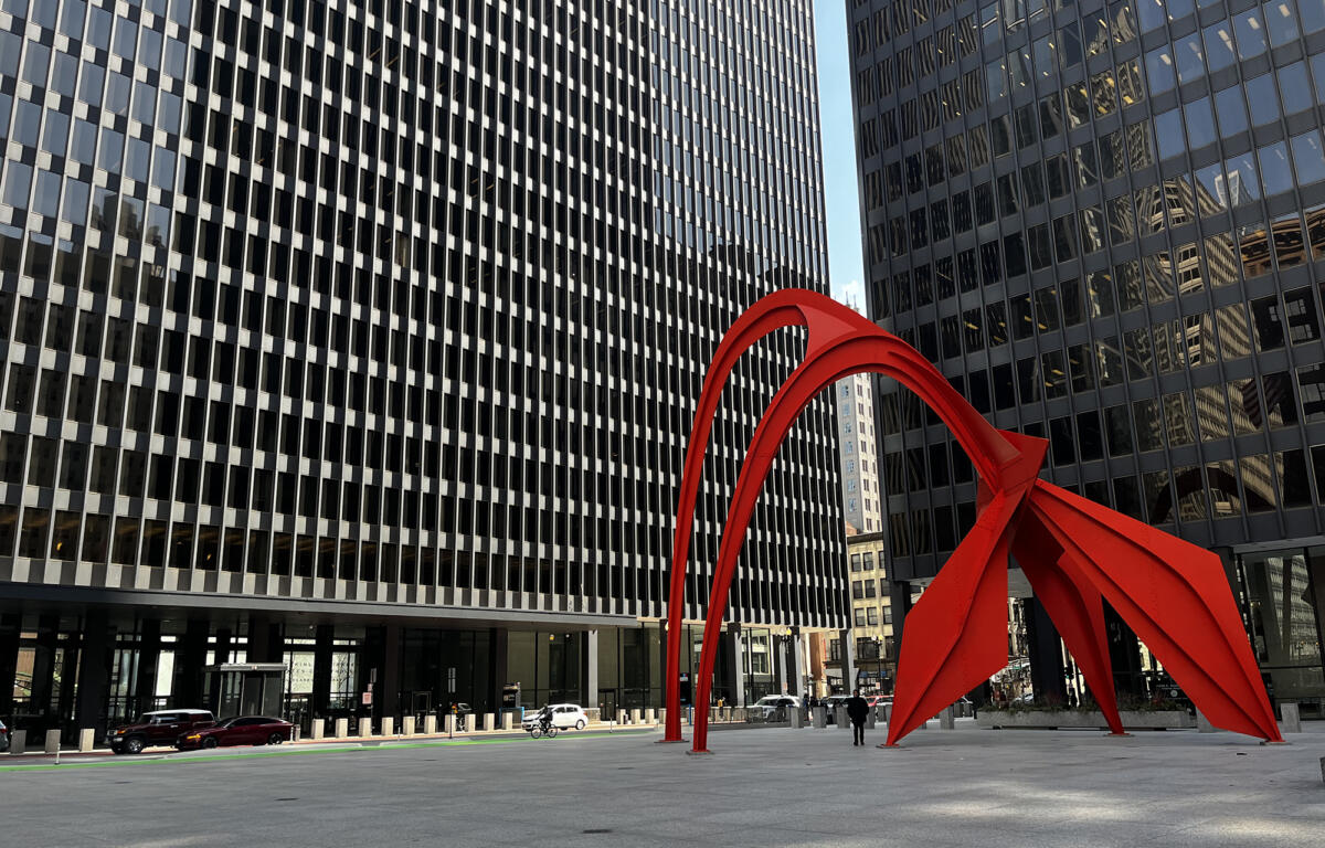Two black glass office towers frame a bright red abstract sculpture in a city plaza you can see cars and pedestrians nearby.