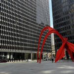Two black glass office towers frame a bright red abstract sculpture in a city plaza you can see cars and pedestrians nearby.