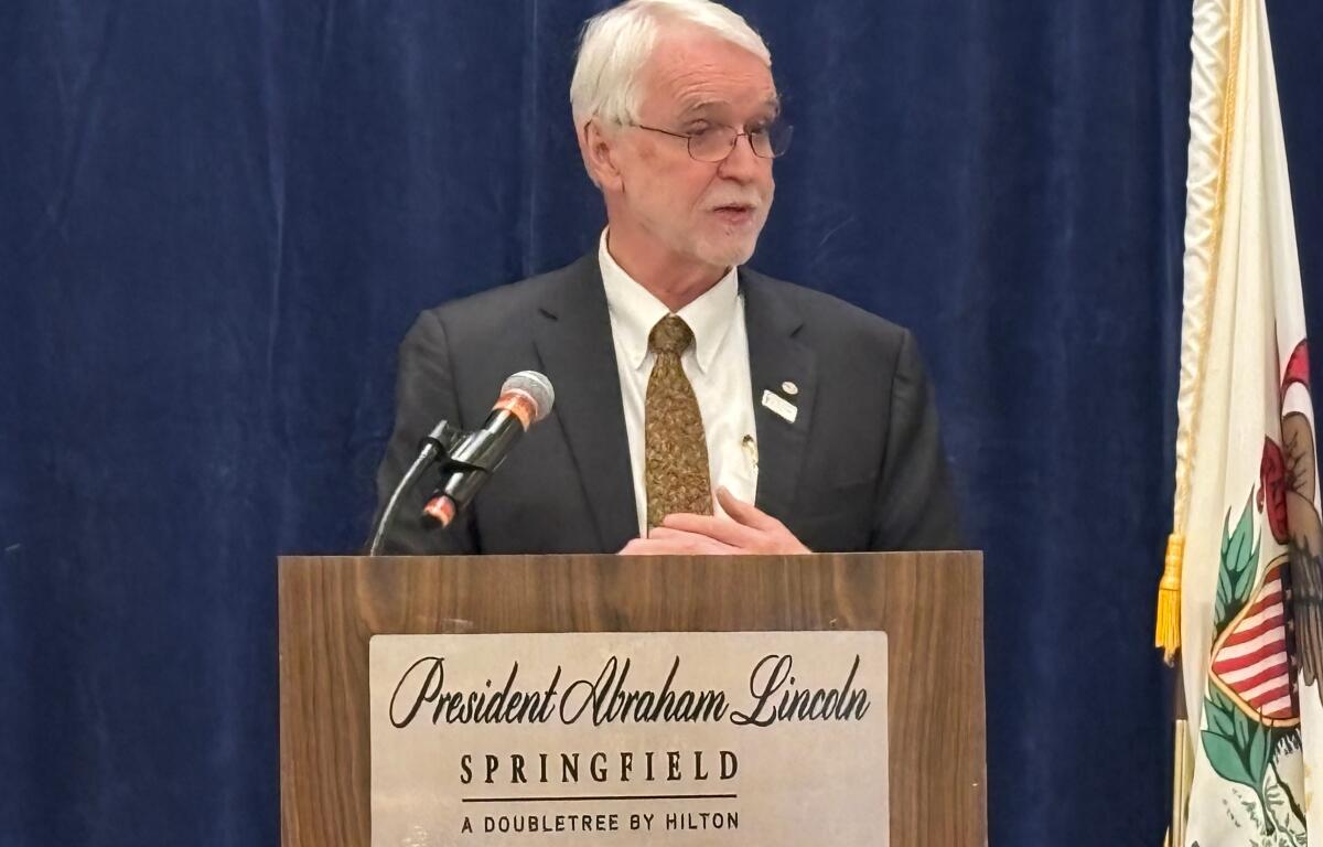 Older man in a suit giving a speech at a lectern labeled 'President Abraham Lincoln, Springfield, A Doubletree by Hilton' against a blue curtain backdrop with a microphone nearby.