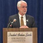 Older man in a suit giving a speech at a lectern labeled 'President Abraham Lincoln, Springfield, A Doubletree by Hilton' against a blue curtain backdrop with a microphone nearby.