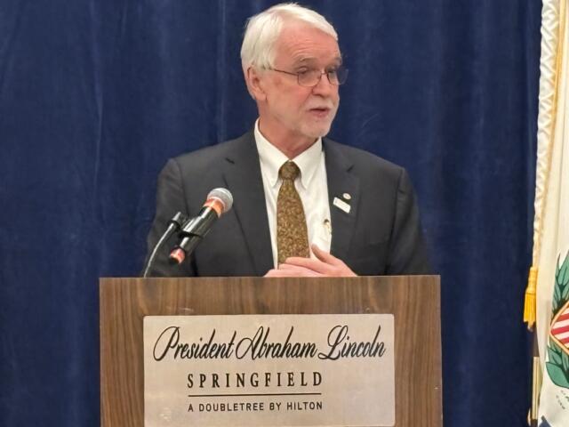 Older man in a suit giving a speech at a lectern labeled 'President Abraham Lincoln, Springfield, A Doubletree by Hilton' against a blue curtain backdrop with a microphone nearby.