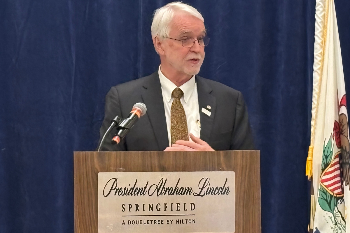 Older man in a suit giving a speech at a lectern labeled 'President Abraham Lincoln, Springfield, A Doubletree by Hilton' against a blue curtain backdrop with a microphone nearby.