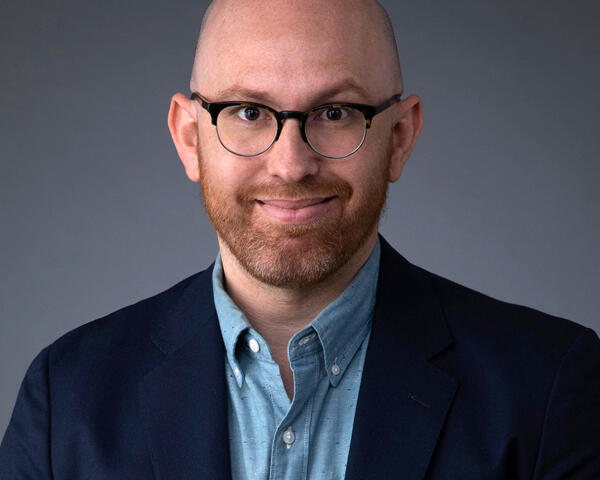 Professional headshot of a smiling, bald man with round glasses, wearing a navy blazer and light blue shirt against a gray backdrop.