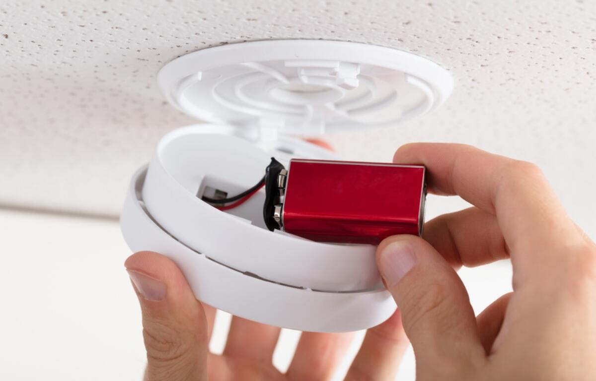 Person inserting a red battery into a white smoke detector housing on the ceiling. When finished, the cover closes back into place.