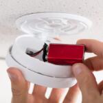Person inserting a red battery into a white smoke detector housing on the ceiling. When finished, the cover closes back into place.